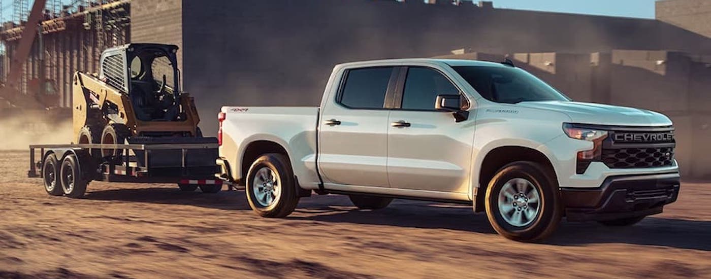 A white 2023 Chevy Silverado 1500 WT is shown from the side while towing a skid-steer.
