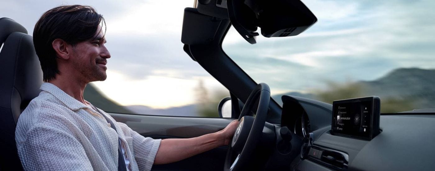 A smiling person is shown driving a 2024 Mazda MX-5 Miata.