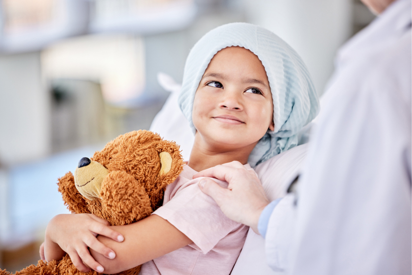Doctor with hand on little girl with cancer holding onto a brown teddy bear