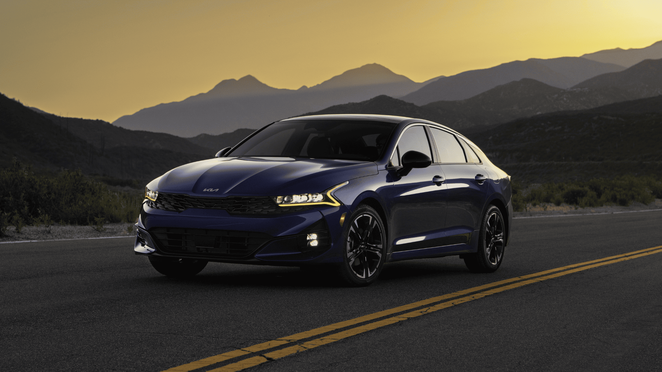 2024 Kia K5 in blue with a sunset backdrop, featuring sleek and bold design.