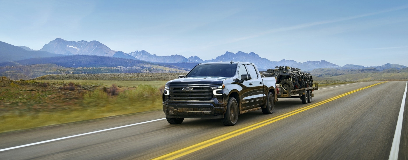 A black 2025 Chevy Silverado 1500 is shown from the front at an angle while towing ATVs.