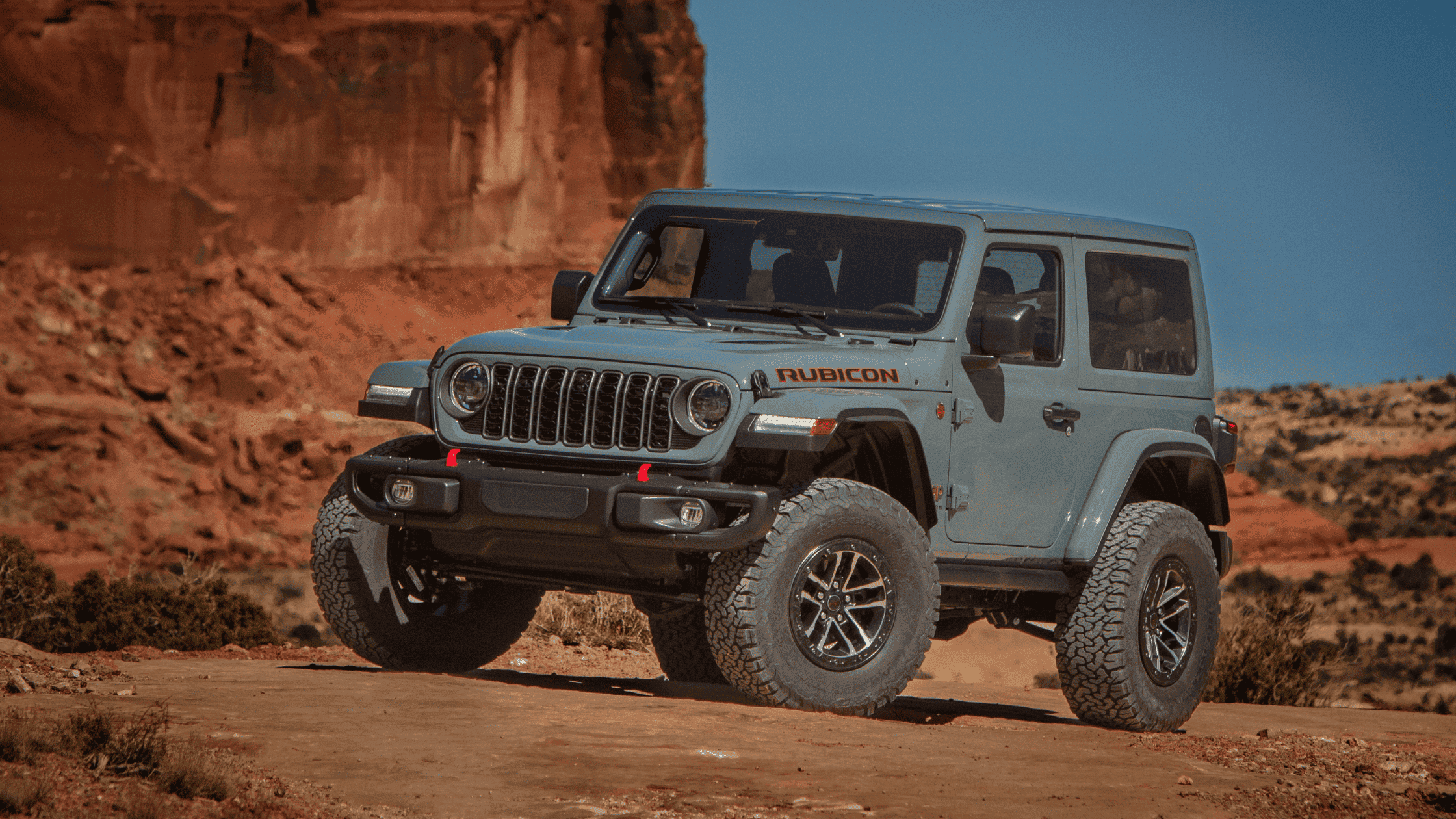 2025 Jeep&reg; Wrangler in gray parked on desert terrain, showcasing front grille, red tow hooks, and off-road tires.