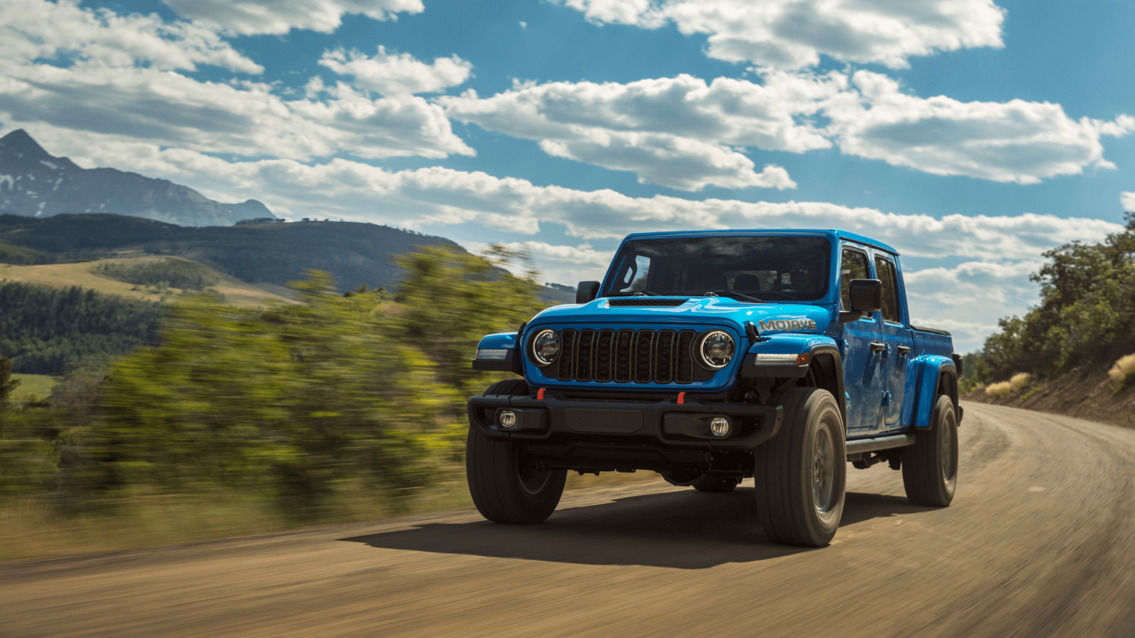2025 Jeep&reg; Gladiator Mojave in Hydro Blue driving on a dirt mountain road, with rugged terrain and scenic sky in the background.