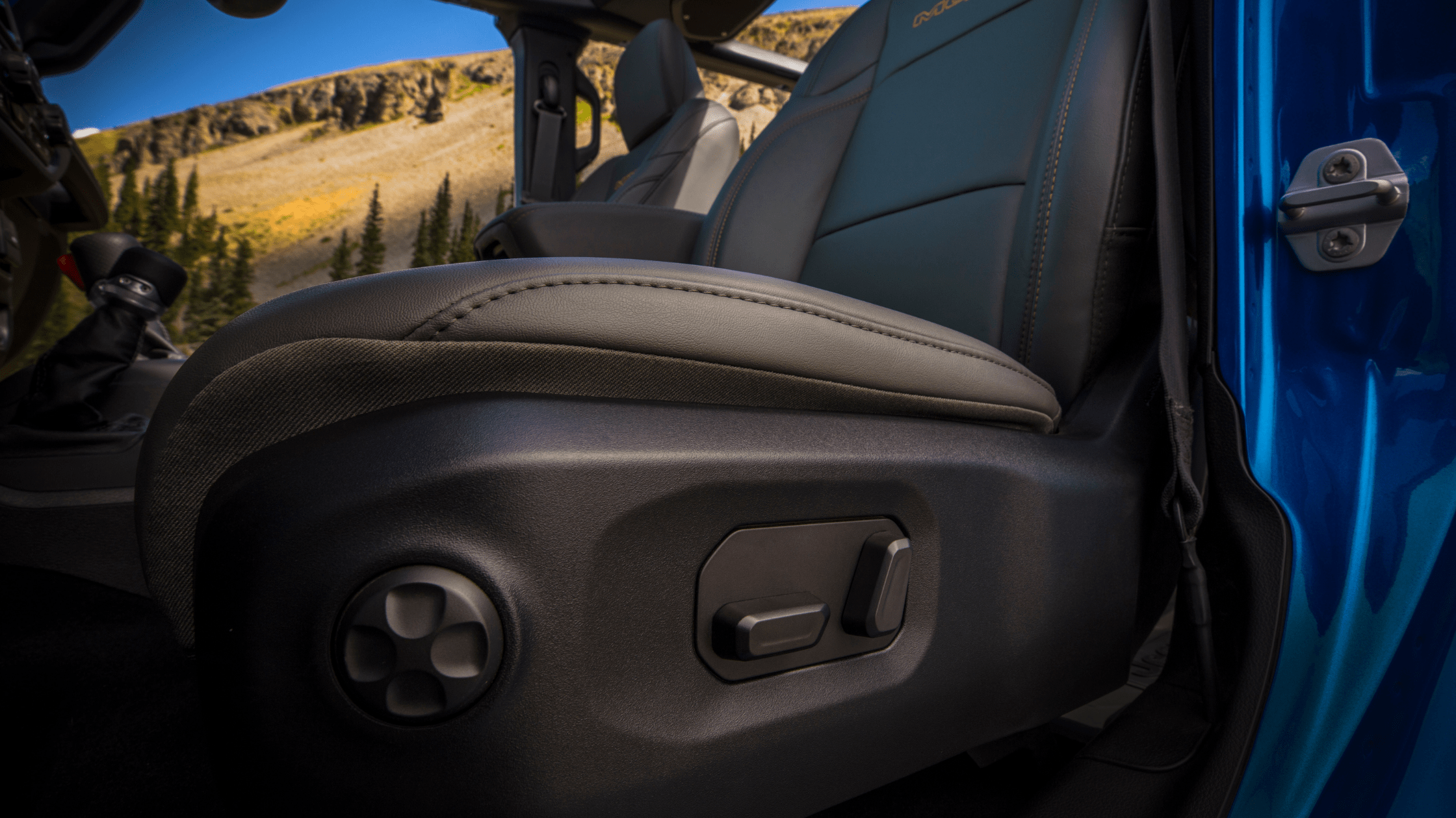 Close-up of the 2025 Jeep&reg; Gladiator's front seat controls with leather upholstery and orange stitching, shown with door open against a mountain background.