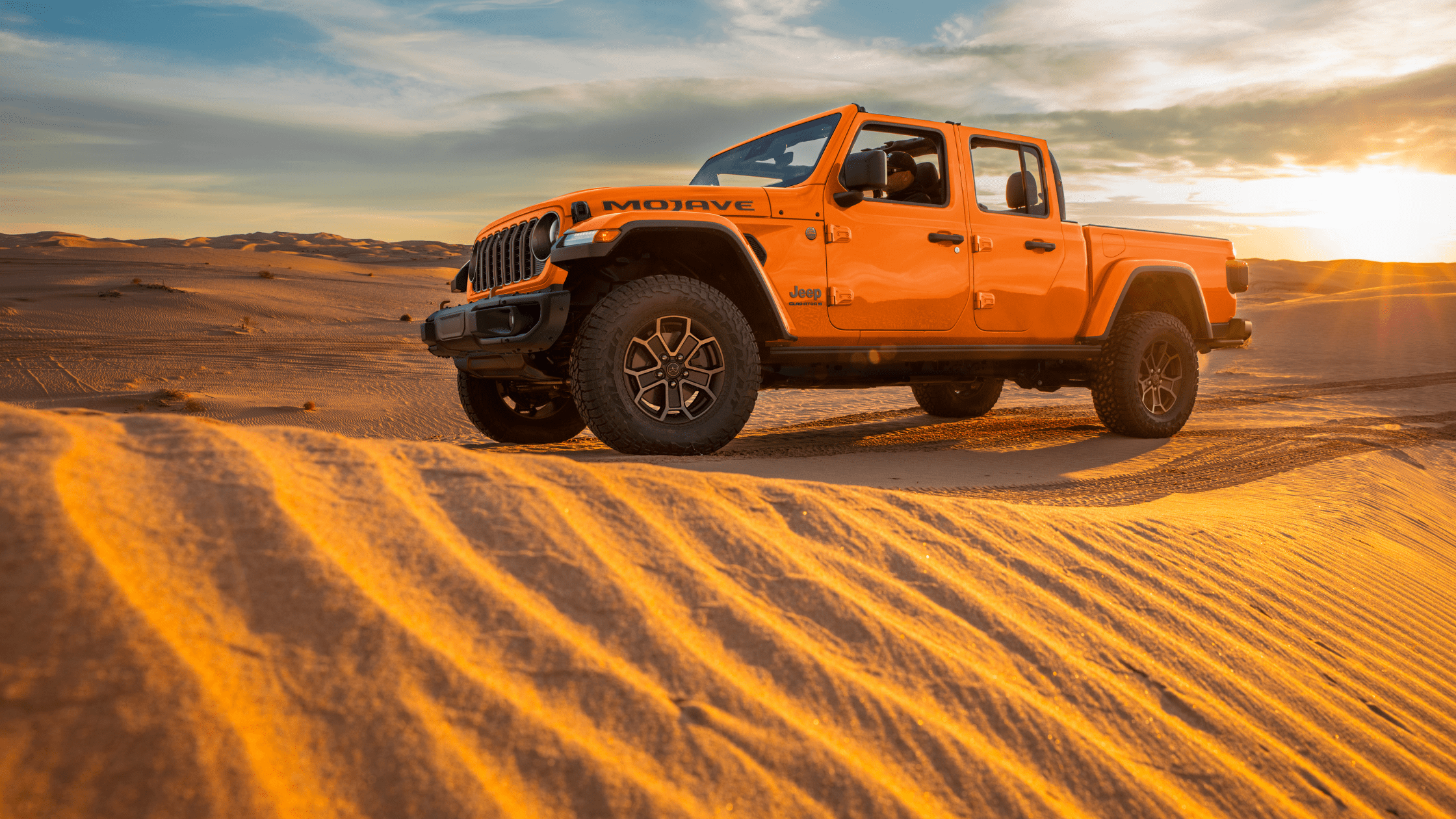 2025 Jeep&reg; Gladiator Mojave in bold orange driving on golden sand dunes at sunset, with open doors and rugged tires.