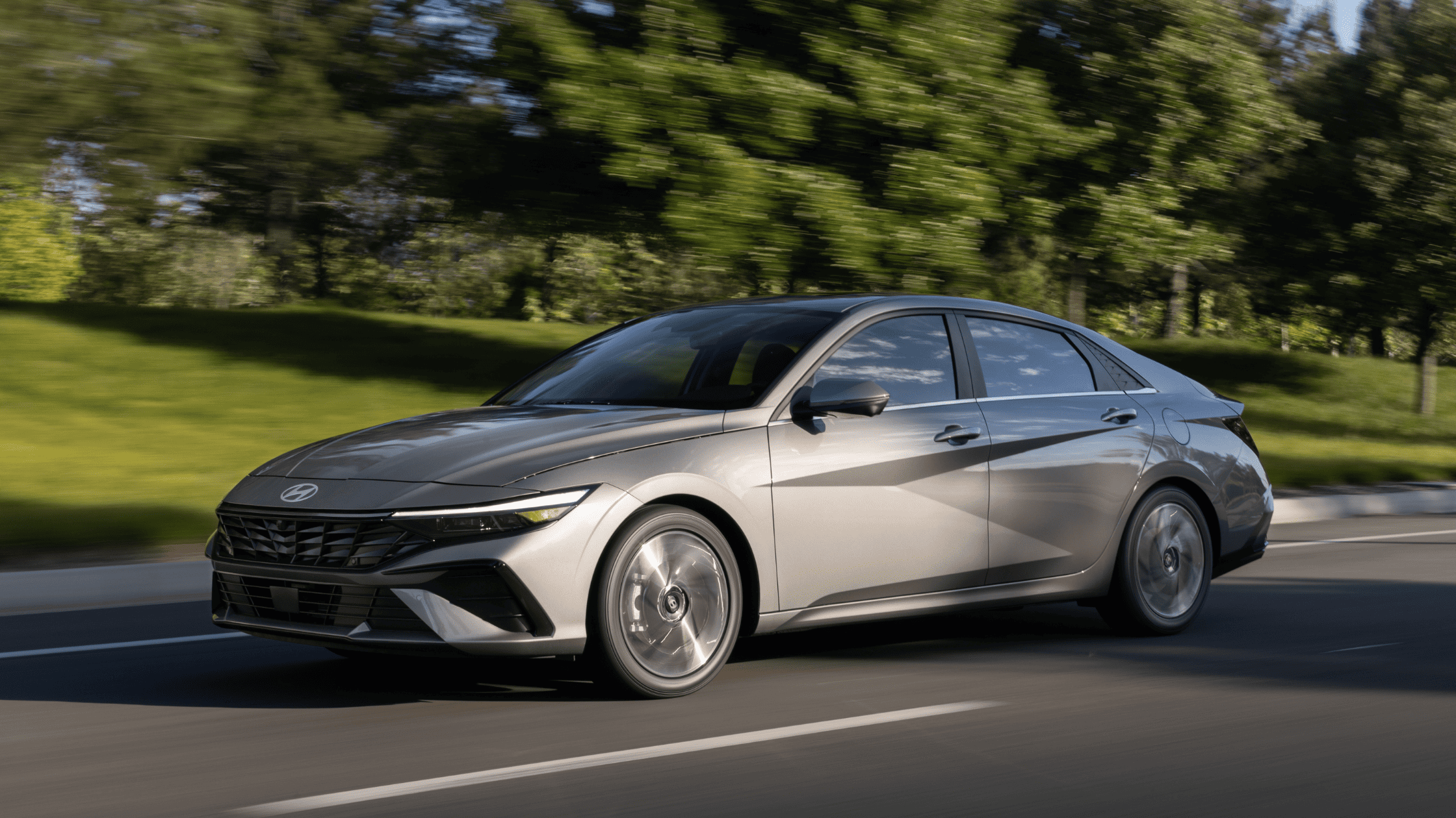 2025 Hyundai Elantra sedan in silver driving on a suburban road with trees in the background