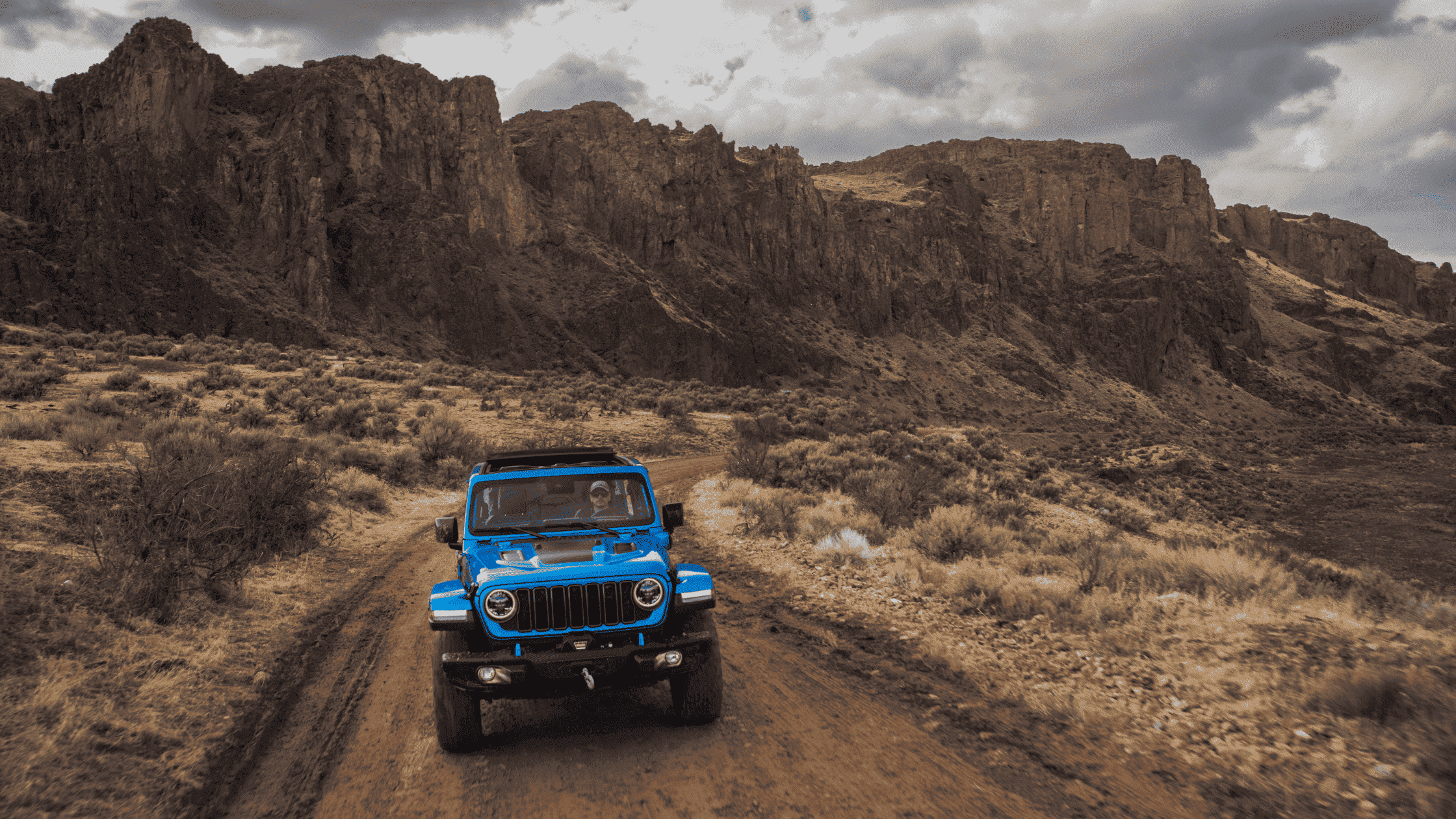 Blue 2025 Jeep Wrangler 4xe driving on a rugged dirt trail through remote canyon landscape