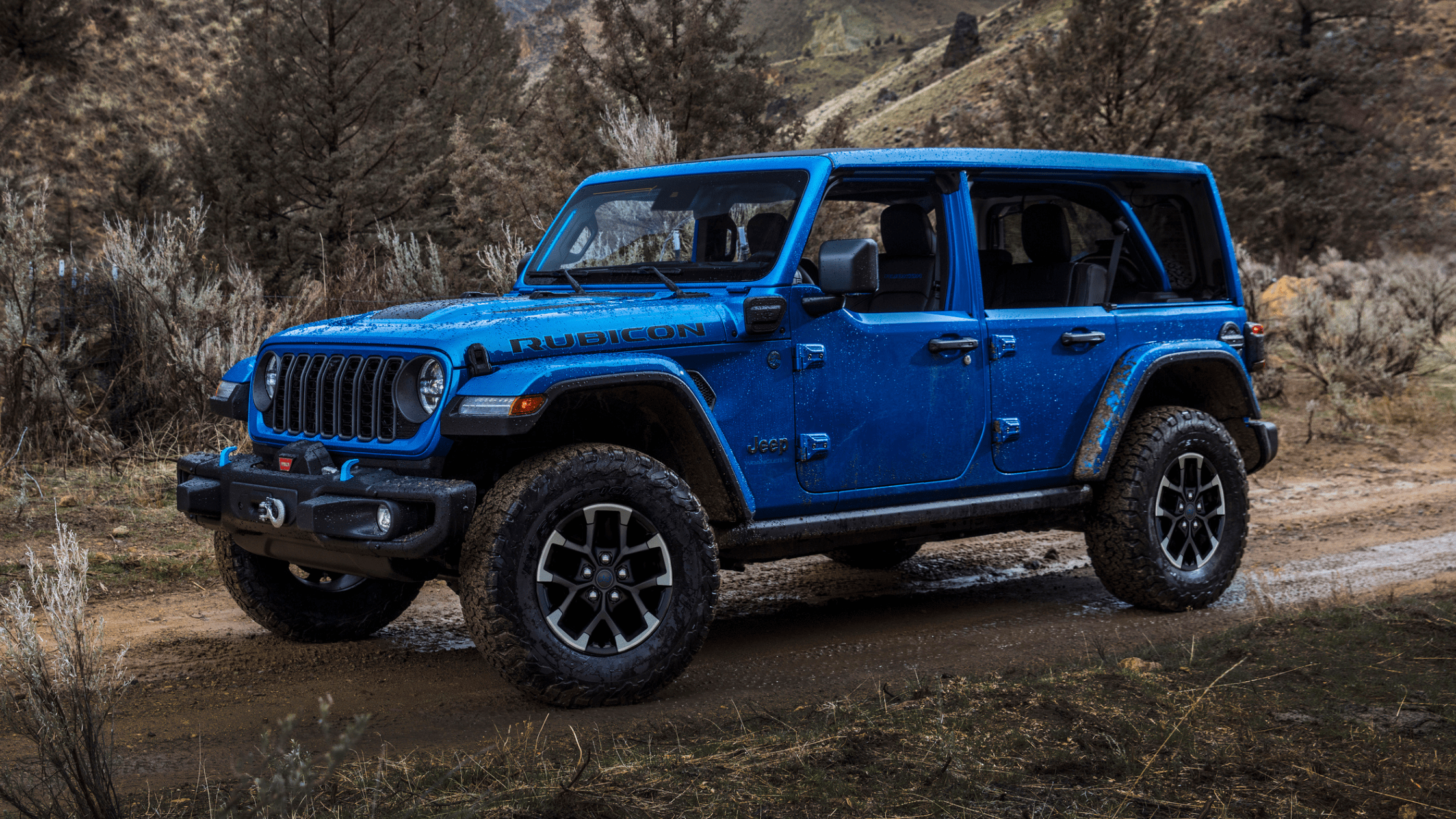 Blue 2025 Jeep&reg; Wrangler 4xe Rubicon parked on a muddy trail with open doors, surrounded by rugged forest terrain.