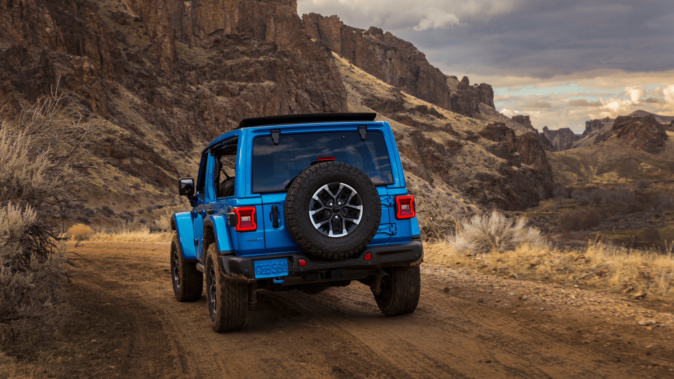 Rear view of a blue Jeep&reg; Wrangler 4xe driving along a dirt trail with rocky mountains in the background.