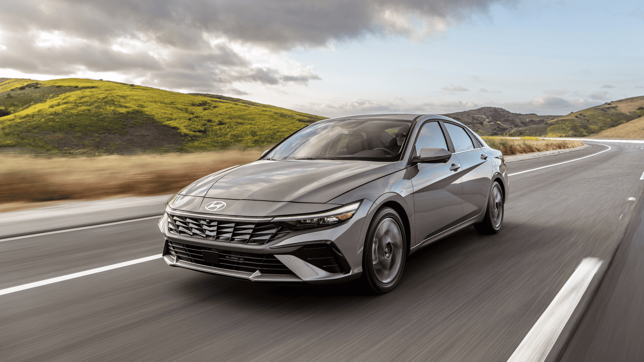 2025 Hyundai Elantra in gray driving on a scenic highway with rolling green hills and a partly cloudy sky in the background.