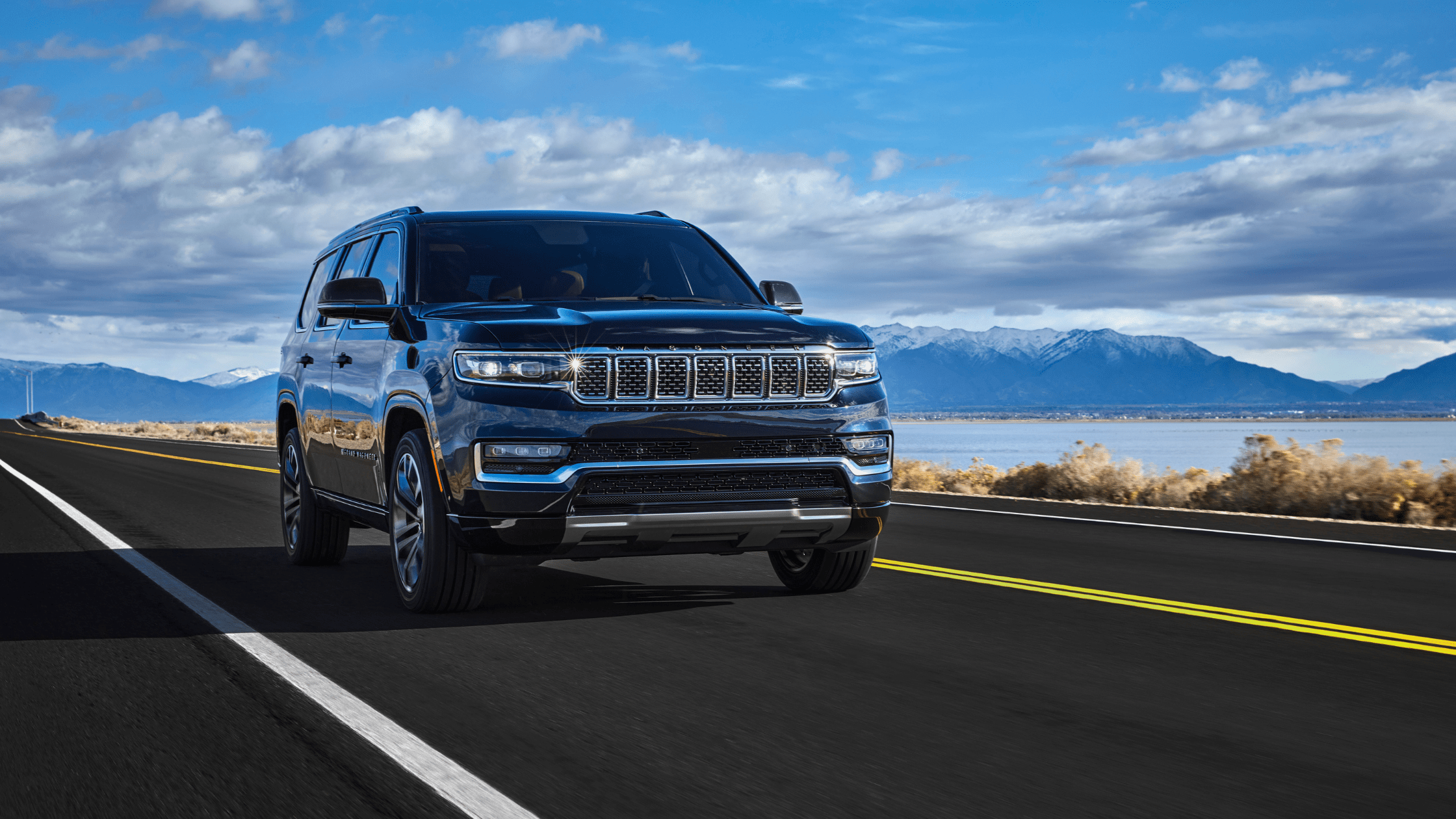2025 Jeep&reg; Grand Wagoneer in dark blue driving along a highway with mountain and lake views.