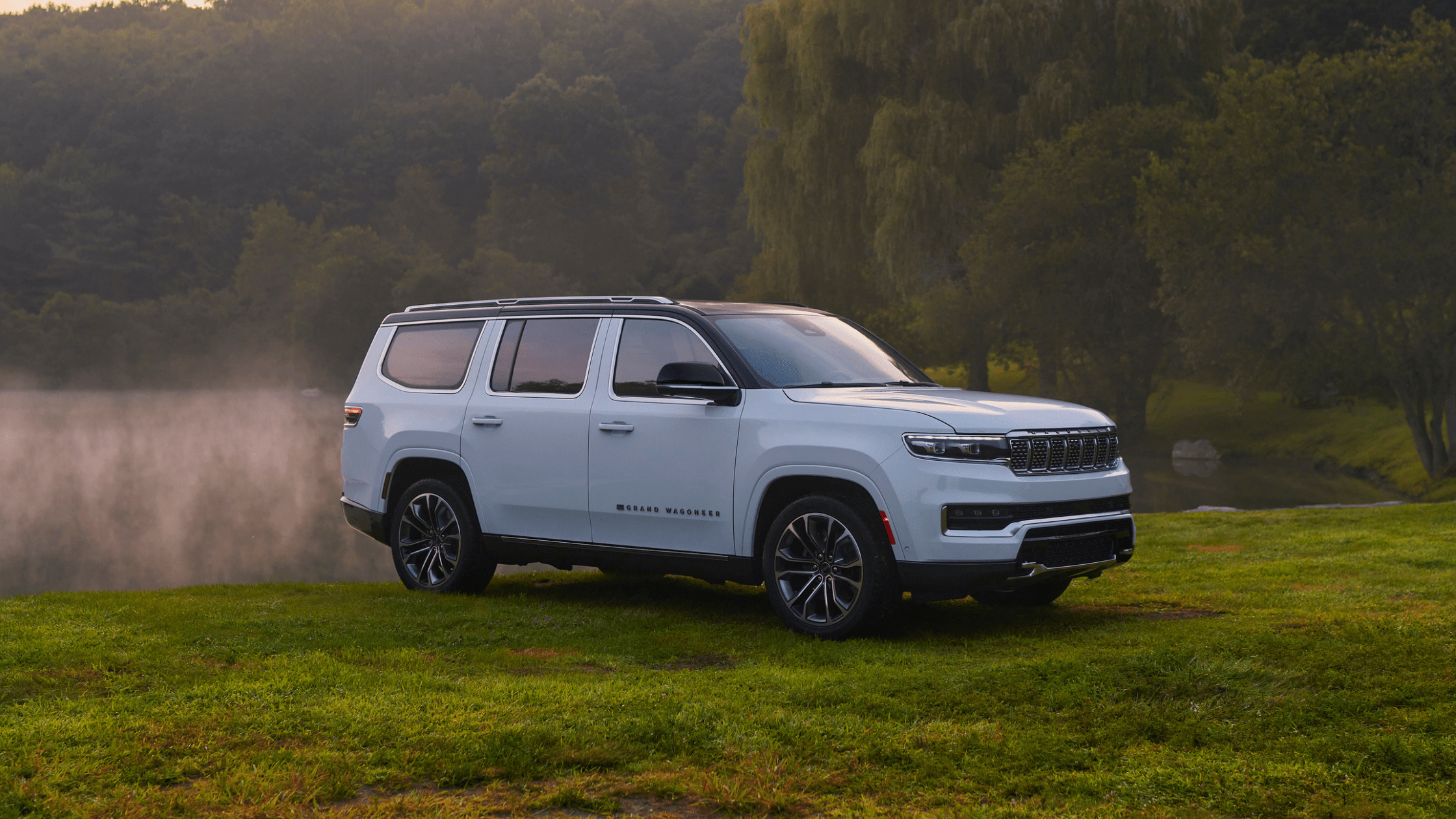 2025 Jeep Grand Wagoneer in white parked on a grassy field near a misty forest pond at sunrise.