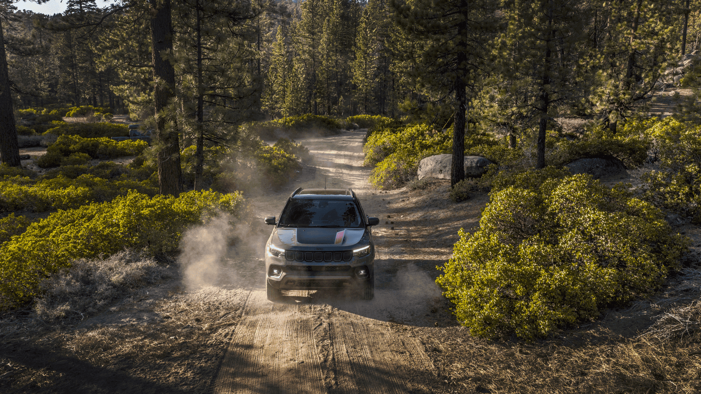 Front view of 2025 Jeep Compass Trailhawk kicking up dust on a forest trail, showcasing off-road capability.