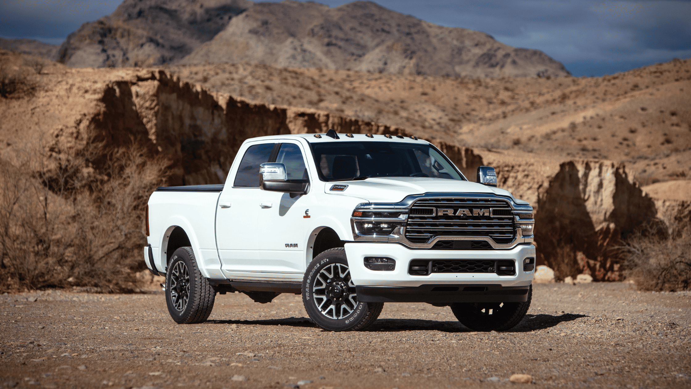 White 2025 Ram 2500 parked in rocky desert terrain, showcasing chrome grille, rugged stance, and off-road tires.