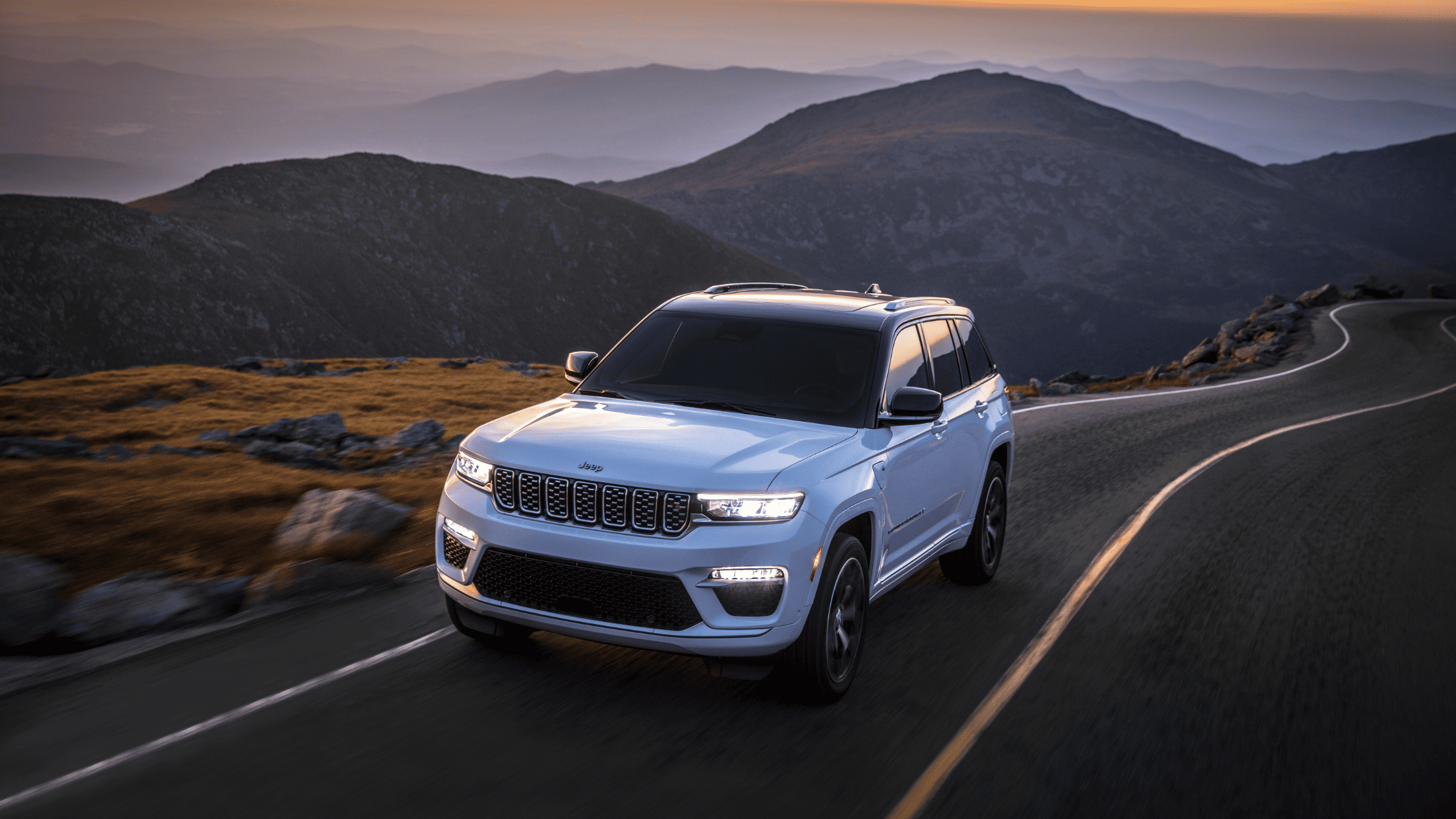 White 2025 Jeep Grand Cherokee 4xe driving on a winding mountain road at sunset, showcasing LED lights and hybrid badge.