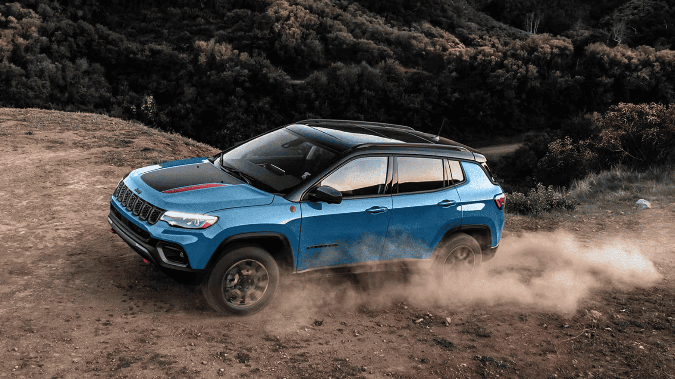 Bright blue 2025 Jeep Compass Trailhawk kicking up dust on a dirt trail, off-road setting with forested hills.