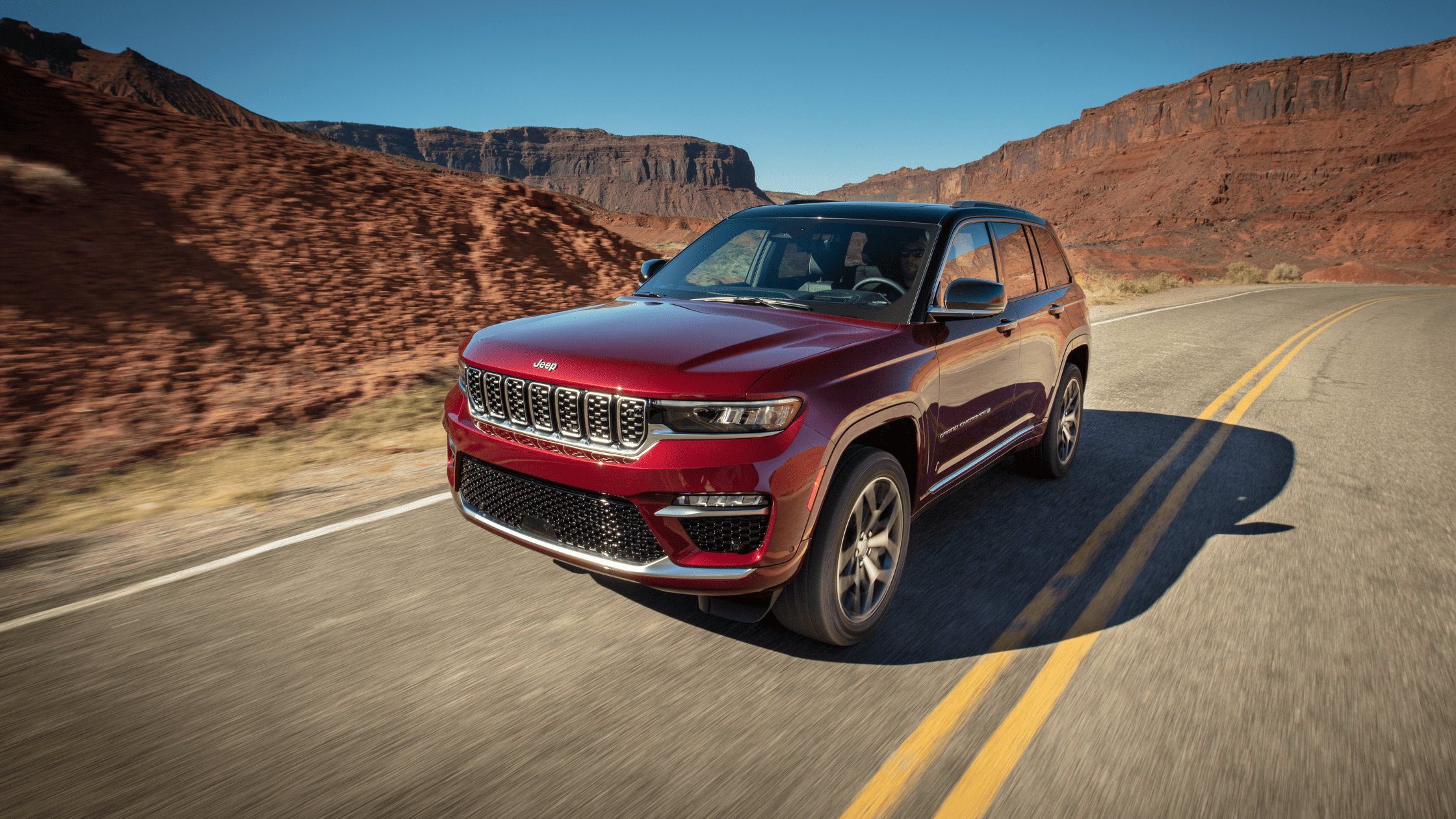 Front-side view of a red 2025 Jeep Grand Cherokee driving through a desert canyon on a winding highway.