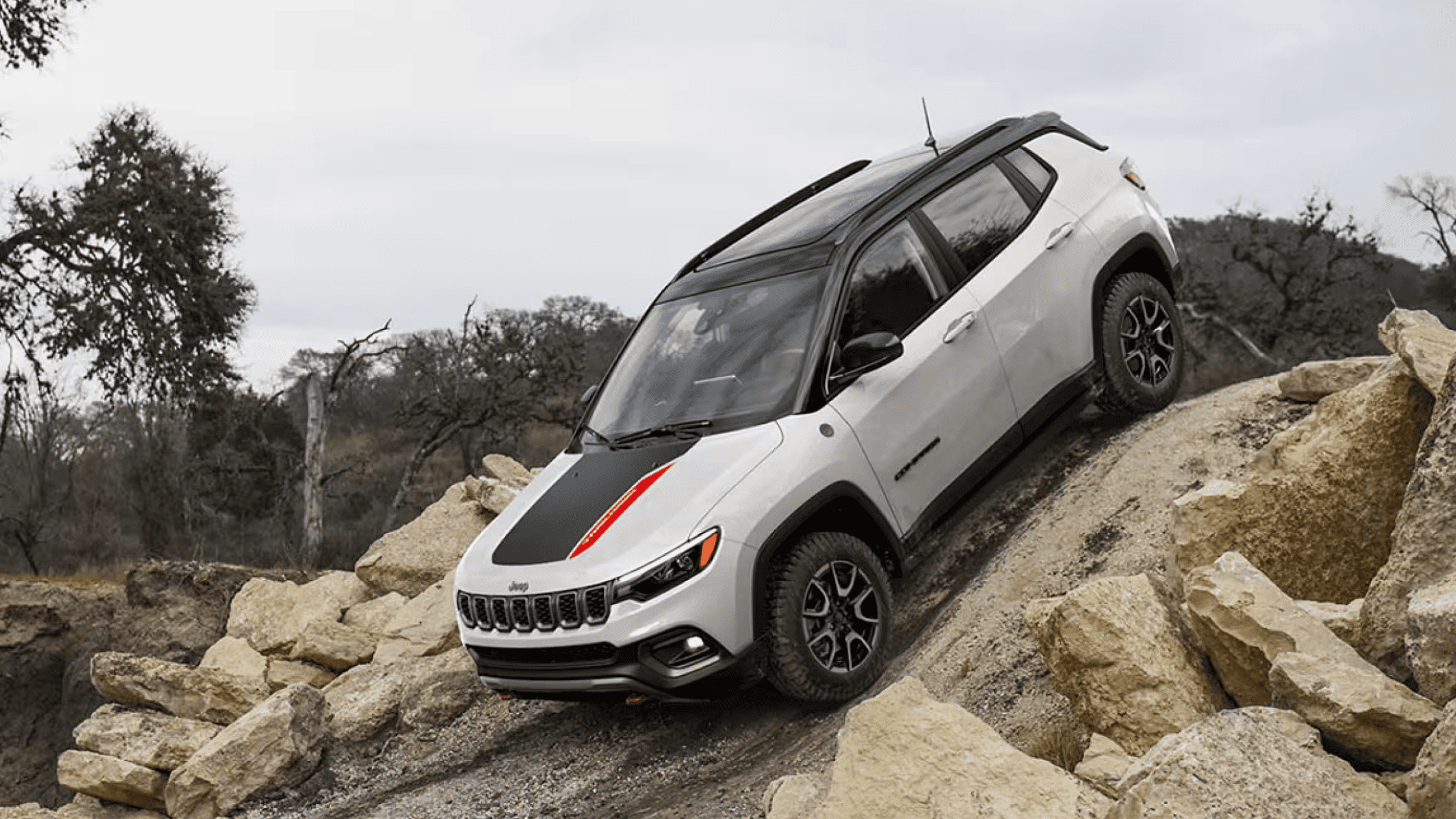 Front-side view of a white 2025 Jeep Compass Trailhawk descending rocky terrain with black and red hood decal.