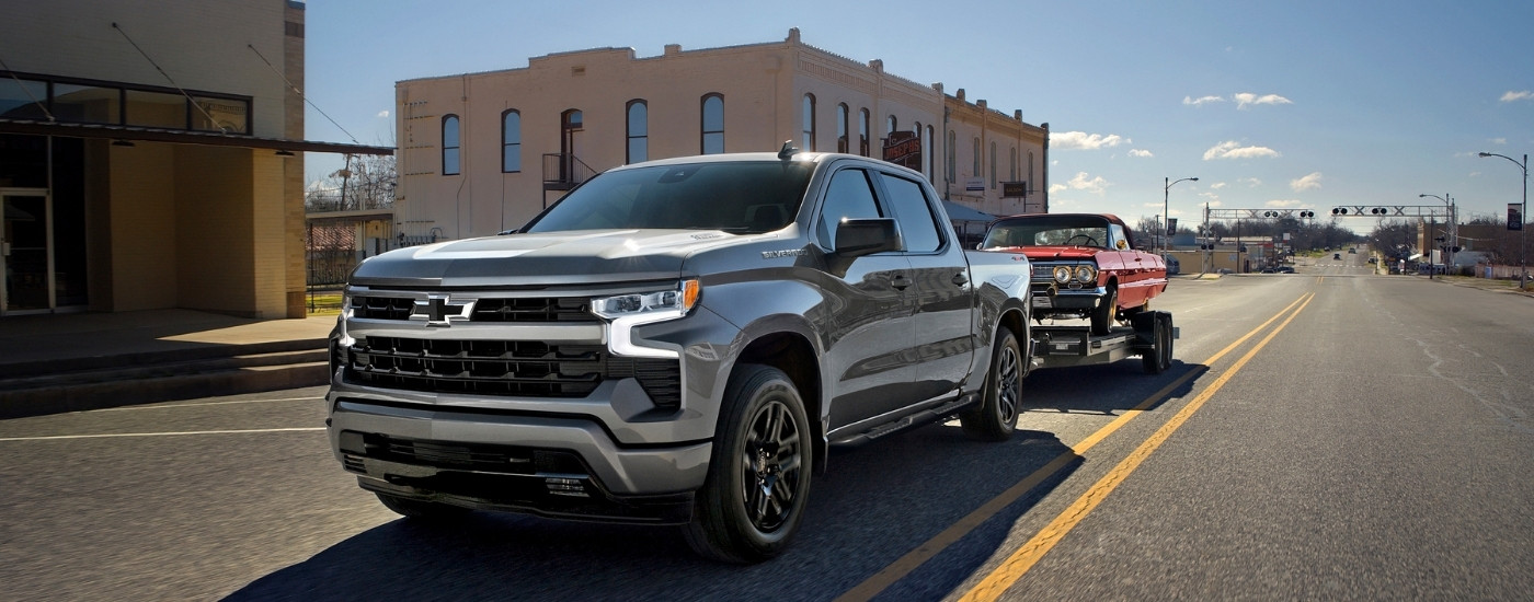 A silver 2025 Chevy Silverado 1500 from the front while towing a red Chevy Impala.