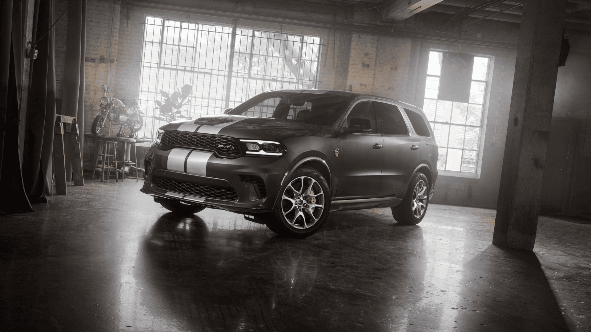 Front angle of a 2026 Dodge Durango SRT Hellcat in matte black with silver racing stripes, parked in a dimly lit industrial garage setting.