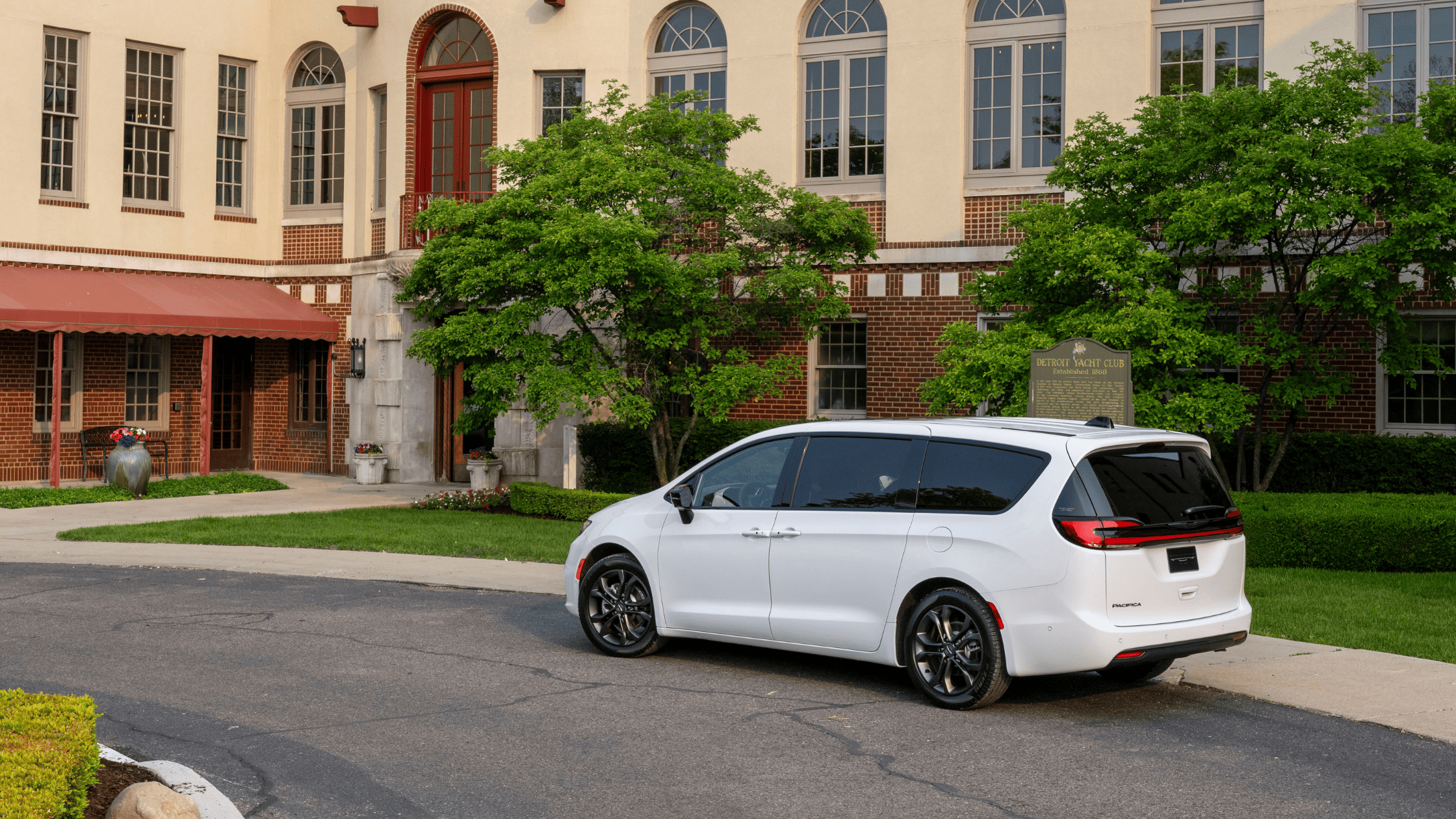 Rear three-quarter view of 2026 Chrysler Pacifica in white parked near a brick building with greenery.