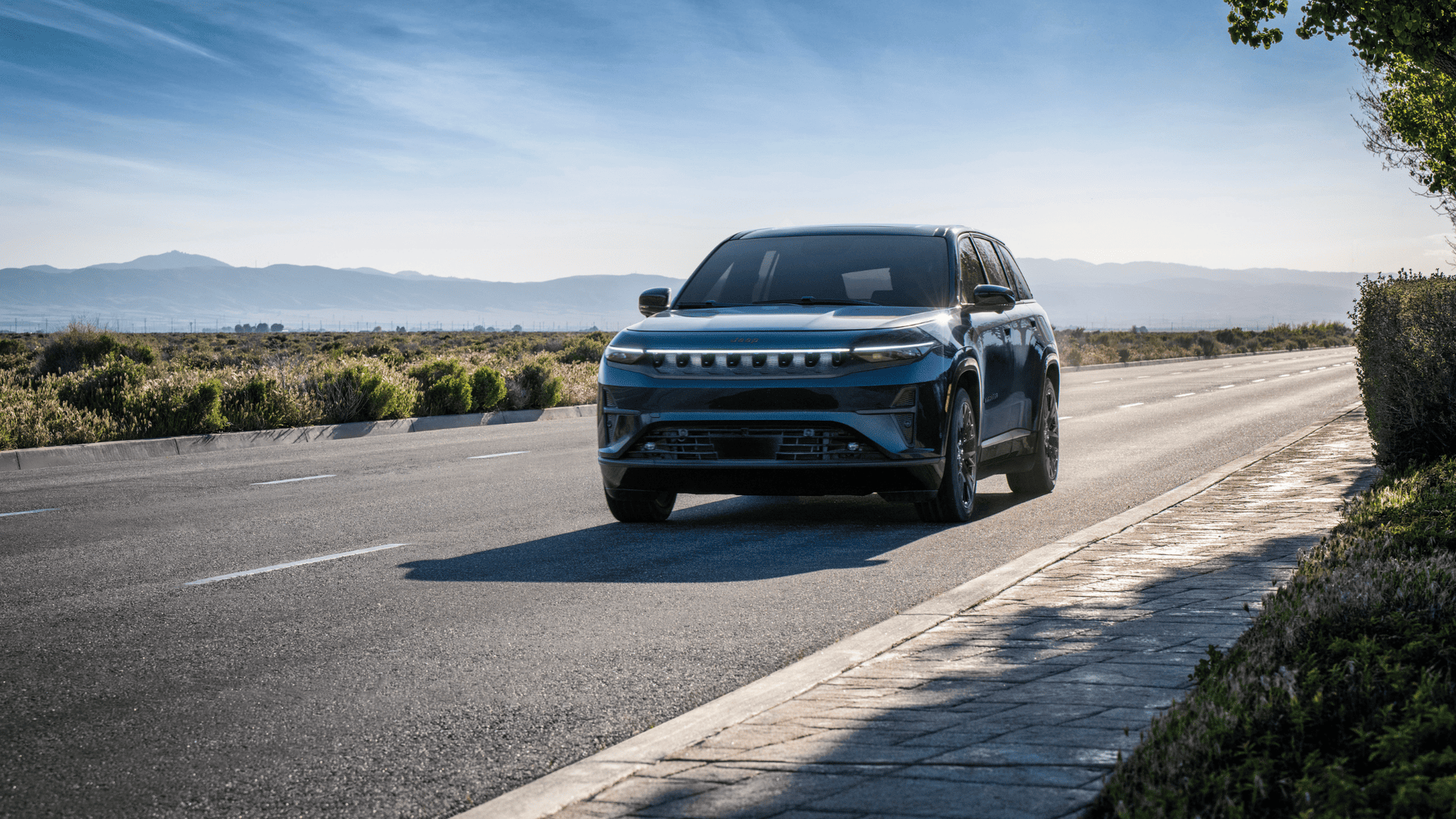 Front view of 2025 Jeep Wagoneer S in dark blue, driving on a sunlit open road with mountains in background.