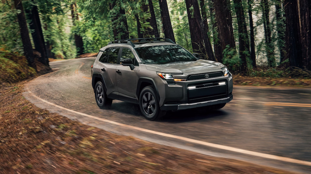 A side view of a taupe 2026 Toyota RAV4 driving down a wooded road