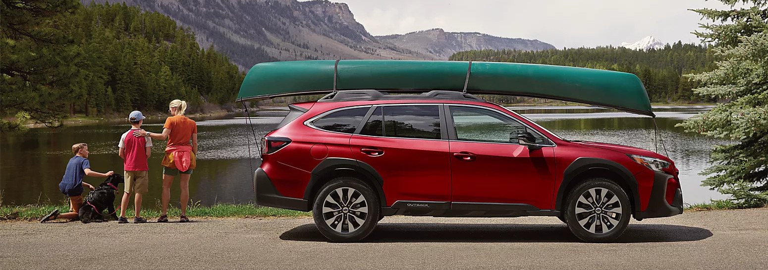 a 2025 subaru outback in red hauling a canoe next to a lake in the mountains with people next to the suv