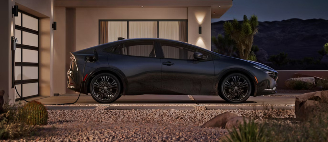 A side-view of a dark-colored 2026 Toyota Prius Plug-In Hybrid charing in a driveway at night