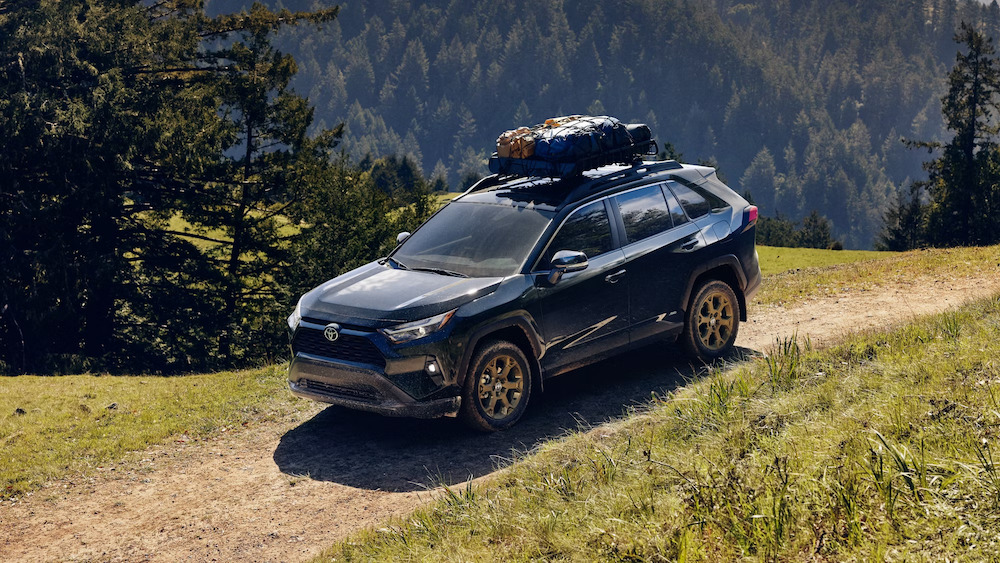 A dark blue 2025 Toyota RAV4 SUV with a packed roof rack driving over a mountain trail
