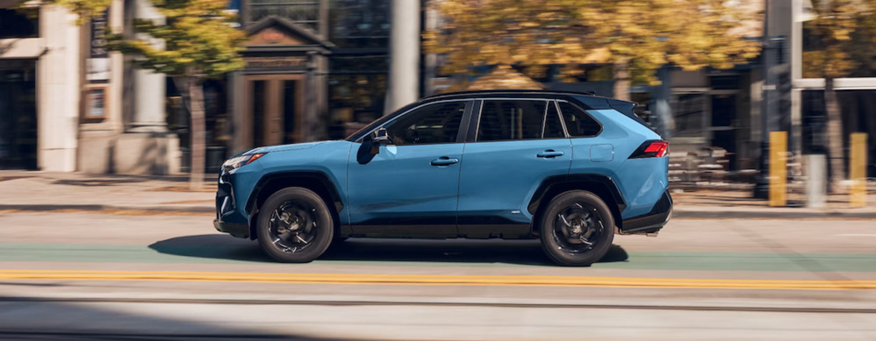 A side view of a blue 2025 Toyota RAV4 Hybrid SUV driving down a city street