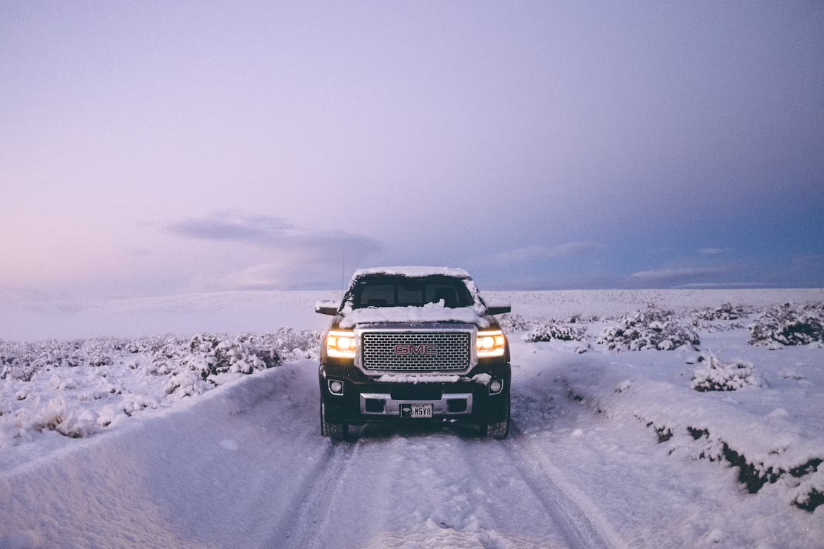 A GMC pickup truck with its headlights on sits on a snowy, desolate road at dusk.