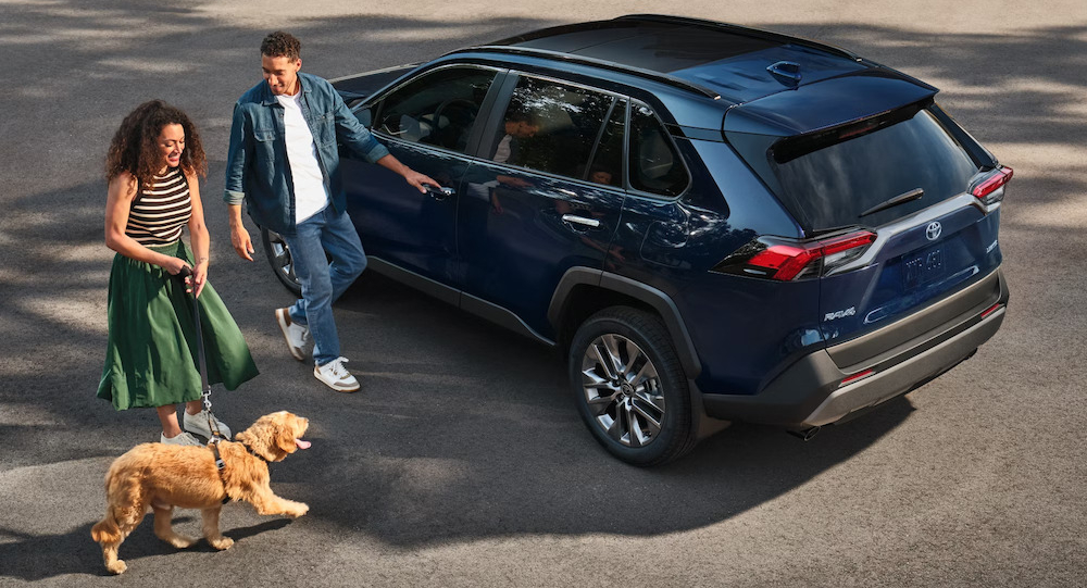 A man and a woman with light brown skin walk a yellow dog beside a dark blue 2025 Toyota RAV4 SUV