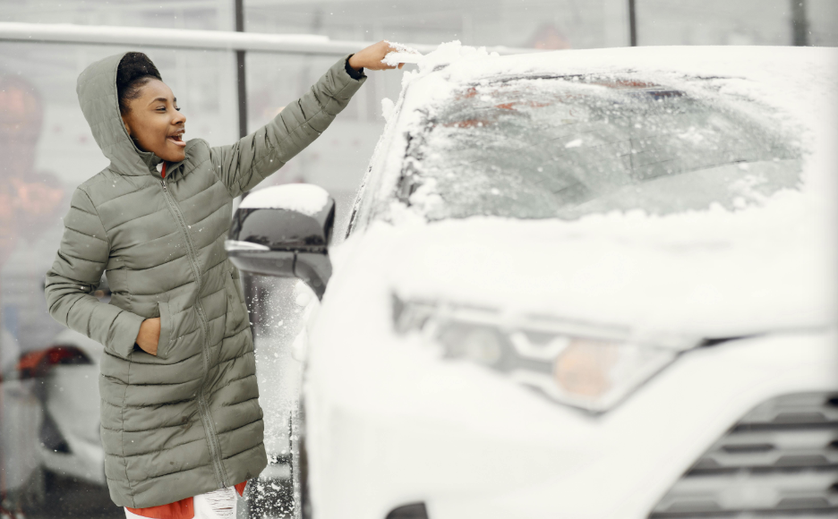 Woman wearing a green winter coat clearing snow off a white SUV windshield