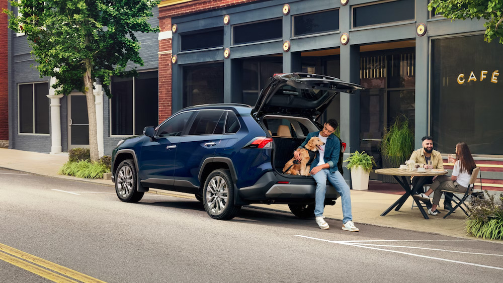 A man with light brown skin sitting with a yellow dog in the edge of the open rear cargo area of a dark blue 2025 Toyota RAV4 SUV