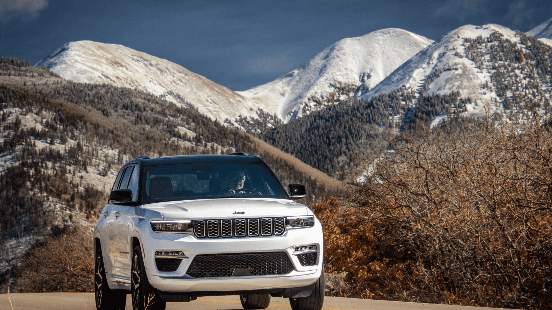 White 2026 Jeep Grand Cherokee driving on a mountain road with snowy peaks in the background.
