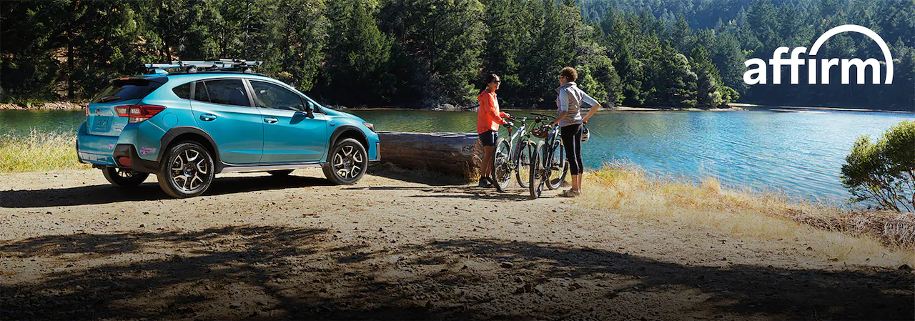 Subaru parked by a lake with roof bike rack.  Two people with bikes nearby.  Affirm logo.