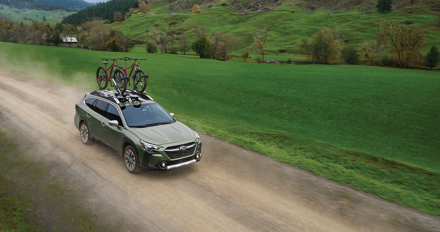 A green Subaru Outback all terrain SUV with two bicycles on its roof rack drives on a gravel road.