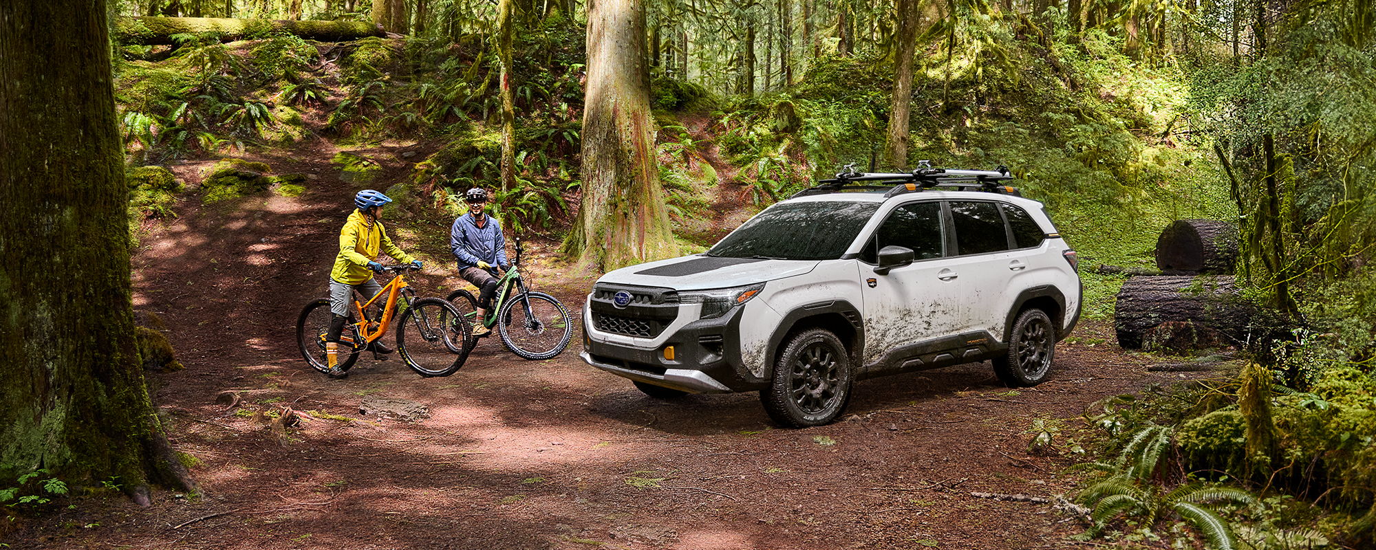 Two people on off-road bicycles next to a muddy 2026 Subaru Forester Wilderness in white in the middle of a forest.