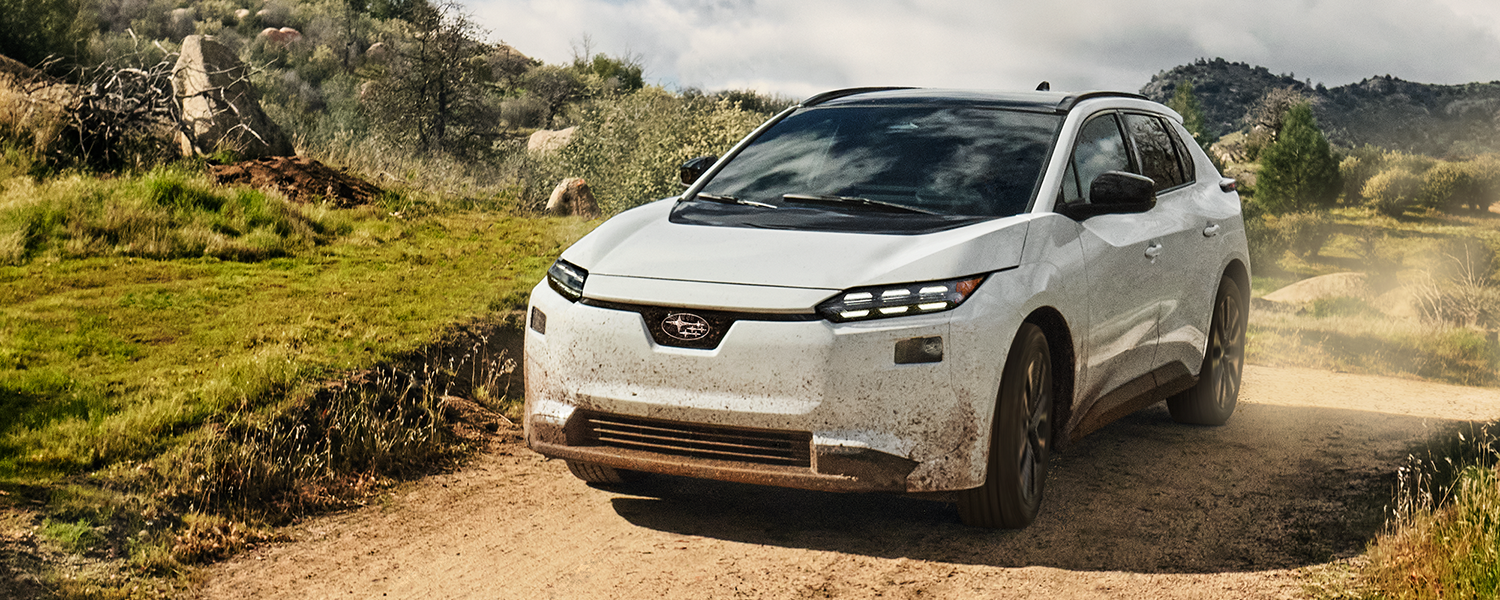 A white 2026 Subaru Solterra electric SUV drives on a dirt road in arid hills, kicking up dust behind.