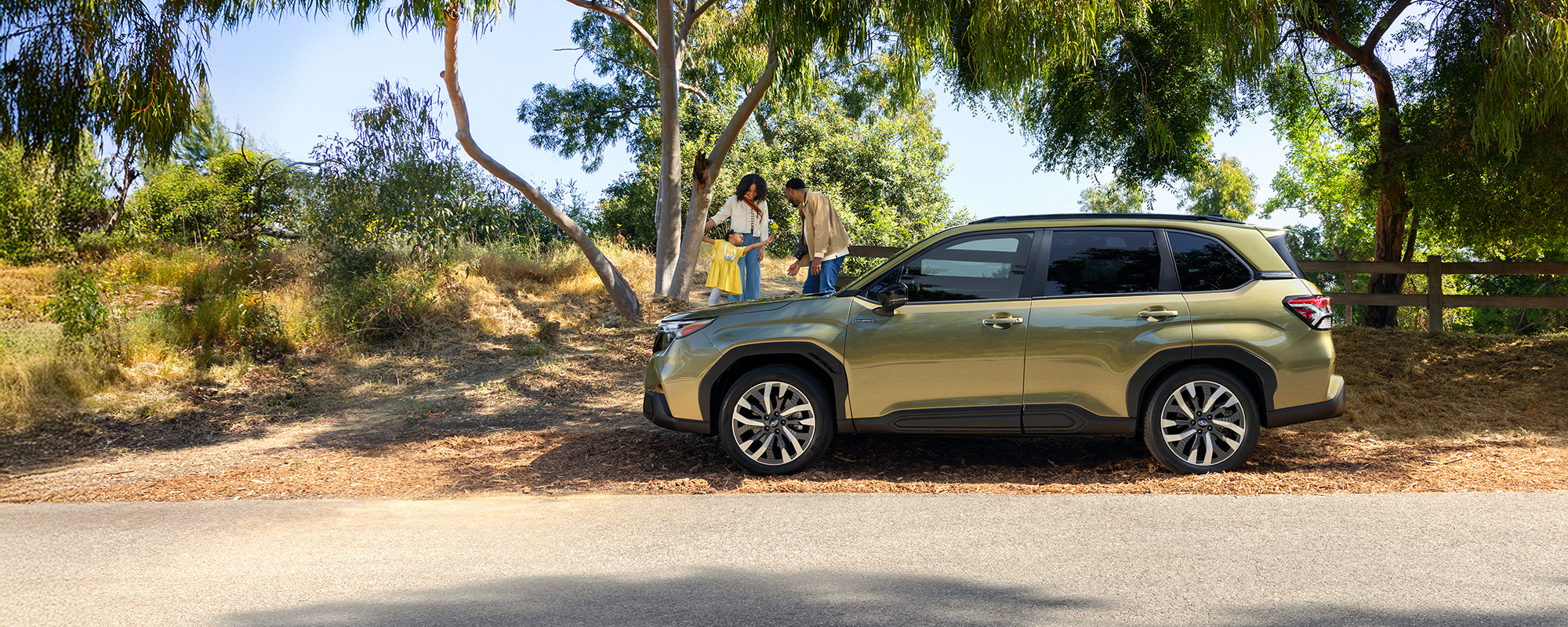 A 2026 Forester Hybrid in Autumn Green Metallic parked on the road as a family plays with their daughter in the background.
