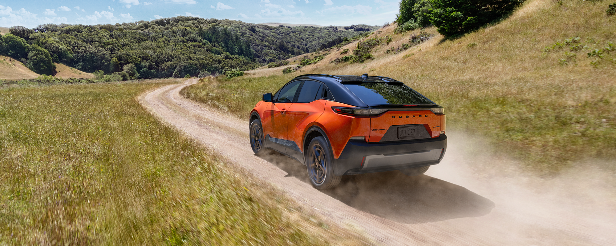 An orange Subaru Uncharted electric SUV drives on a dirt road through some rolling hills.
