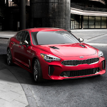 2019kiastingerincoloradospringsatphillongkia The front angle of a shiny red 2019 Kia Stinger as it makes a right turn on an empty city street in front of a big industrial complex