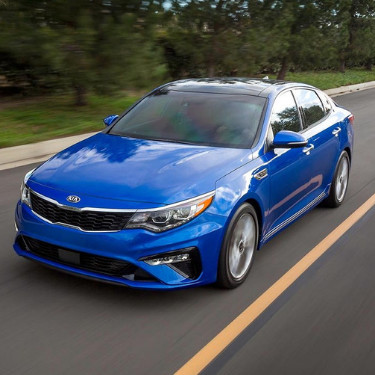 2019kiaoptimaincoloradospringsatphillongkia An angle of the front driver side of a shiny blue 2019 Kia Optima as it speeds down an open road with green trees lining the road